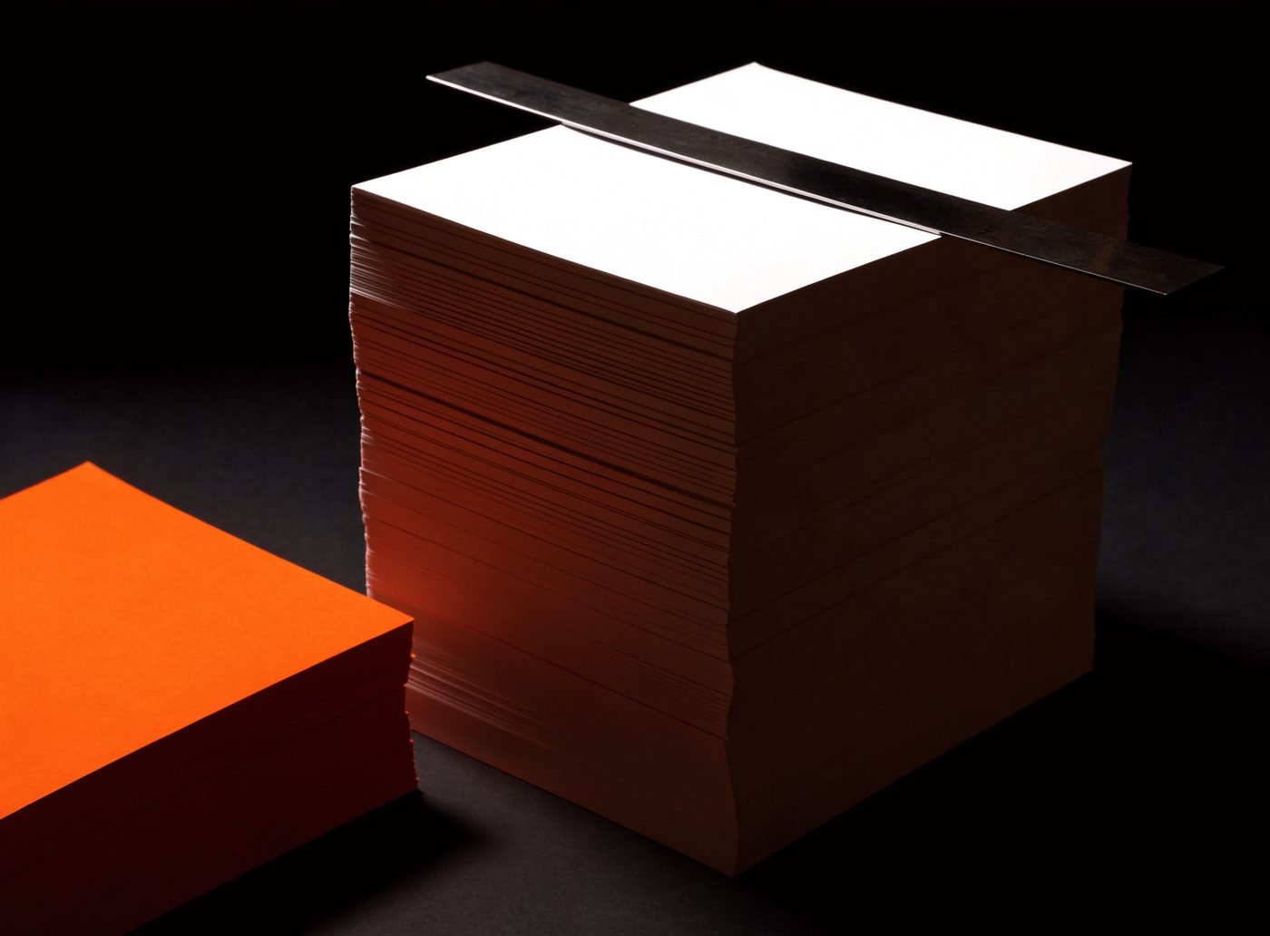 A tall stack of blank white printed sheets with crisp cut edges beside a block of orange stock and a steel straightedge, on a dark surface