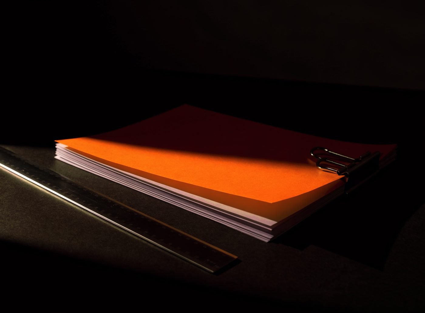 A stack of blank print proofs held by a bulldog clip with an orange sheet on top, beside a steel ruler on a dark surface