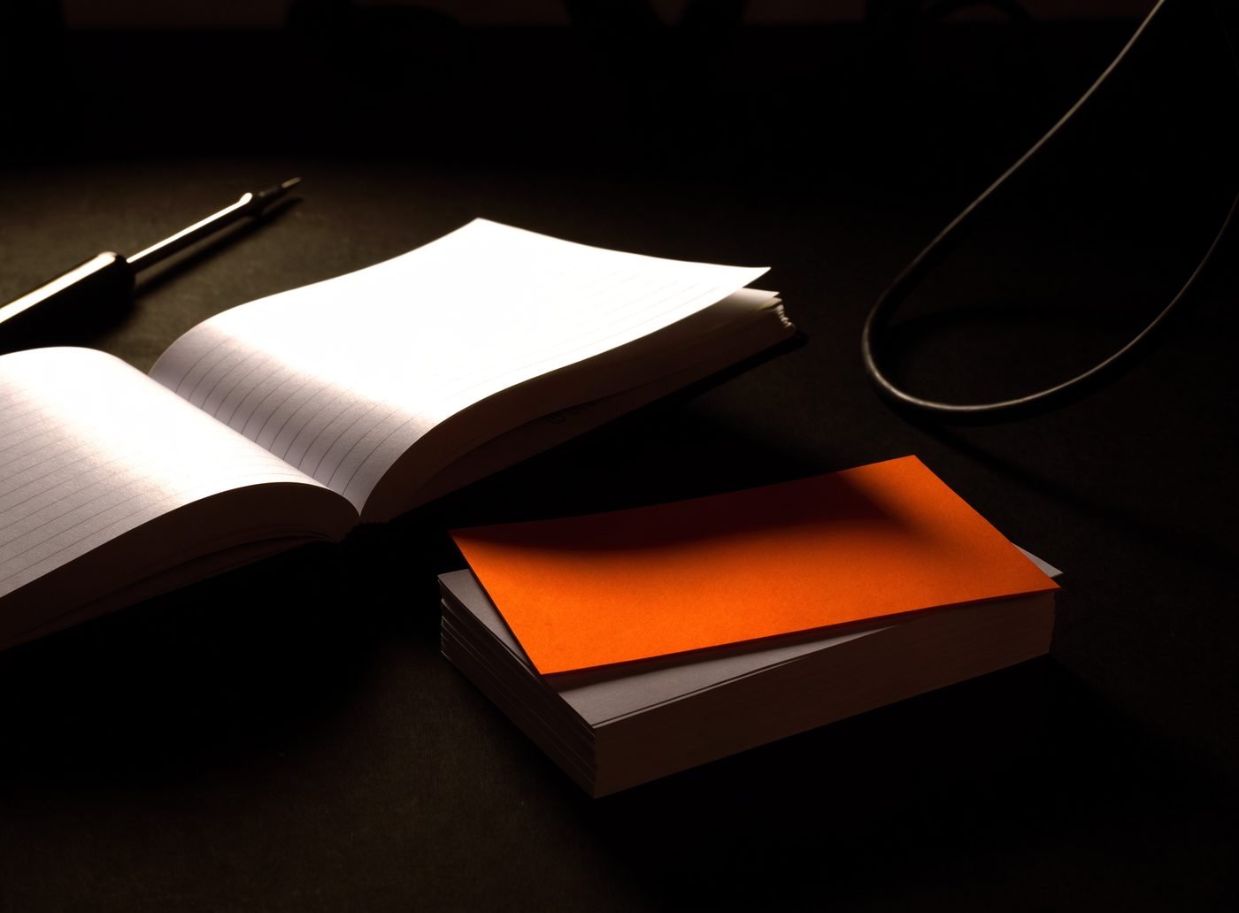 An open blank notebook, a pen, a coiled cable and a stack of blank cards topped with an orange sheet, on a dark bench