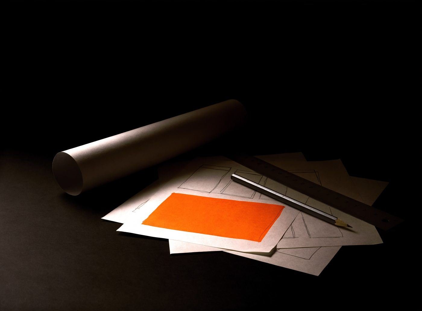 Hand-drawn website wireframe sketches on a dark studio surface, one sheet marked with an orange block, beside a pencil and a rolled paper tube